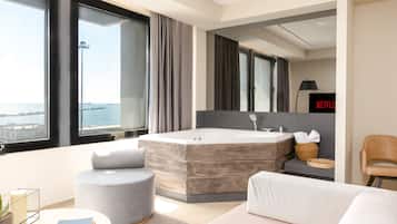 A modern hotel room with a large bathtub, a sofa, a round coffee table, and a view of the sea.