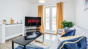 Ein modernes Wohnzimmer mit einem Flachbildfernseher, der „NETFLIX“ anzeigt, einer weißen Ablage mit Dekoration, einer Couch mit Kissen und einem großen Fenster mit Vorhängen.