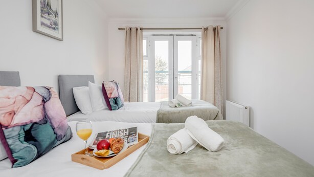 Ein Hotelzimmer mit zwei Betten, einem Frühstückswagen mit Obst und einer Zeitschrift, und Blick auf ein Gebäude durch das Fenster.