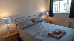 A neatly made bed with a grey duvet, two bedside tables with lamps, a window with curtains, and a radiator.
