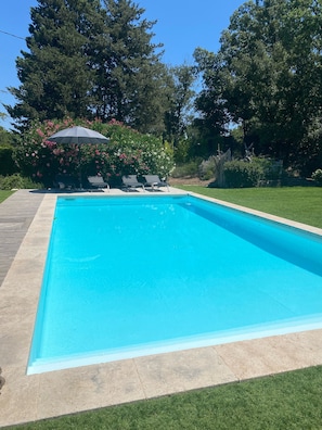 A rectangular swimming pool with a clear blue water surface, surrounded by a concrete deck. There are two lounge chairs and a patio umbrella in the background.