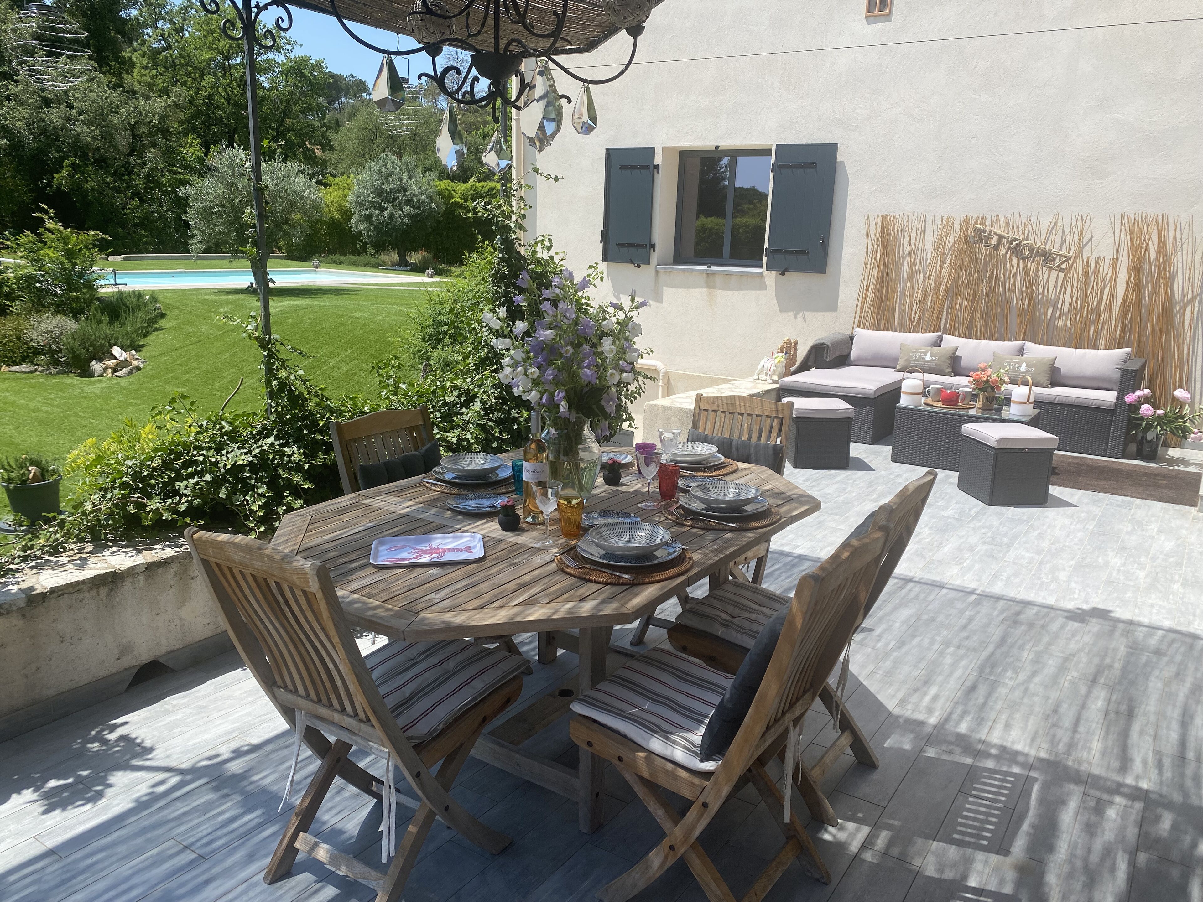 Une terrasse extérieure avec une table à manger pour quatre personnes, entourée de meubles en osier et une piscine en arrière-plan.