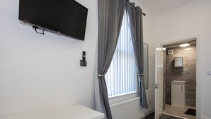 Habitación de hotel con televisor montado en la pared, una ventana con cortinas grises, un radiador blanco, un lavamanos pequeño y una puerta que da a otra habitación.