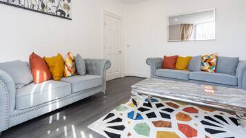 A modern living room with two grey sofas, colorful cushions, a geometric rug, a marble coffee table, a large abstract painting, and a mirror on the wall.