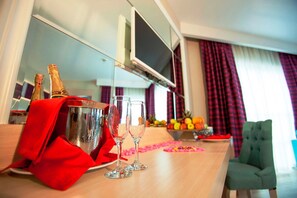 Une chambre d’hôtel équipée d’un mini-bar, de deux verres, d’une bouteille de champagne, d’un panier de fruits et d’une télévision.