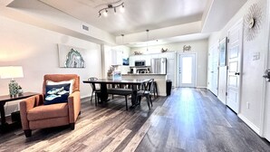 A spacious living room with a dark wood floor, a brown armchair with a fish-patterned pillow, a dining table with chairs, and a kitchen area with white cabinets and stainless steel appliances. - Condo w/3 Rooms, Heated Pool, and Close to Beach! (Corpus Christi)