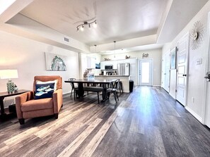 Amplia sala de estar con piso de madera oscura, un sillón marrón con un cojín estampado con motivos de peces, una mesa de comedor con sillas y un área de cocina con gabinetes blancos y electrodomésticos de acero inoxidable.