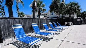 A row of blue lounge chairs with metal frames, arranged on a concrete surface, with a wooden fence and palm trees in the background. - Condo w/3 Rooms, Heated Pool, and Close to Beach! (Corpus Christi)