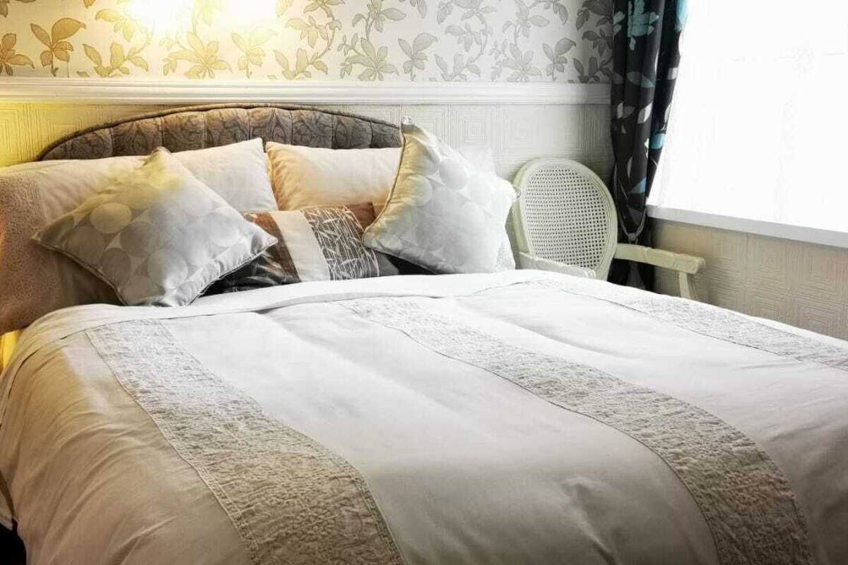 A neatly made bed with a patterned bedspread and pillows, a bedside lamp, and a chair near a window with curtains.