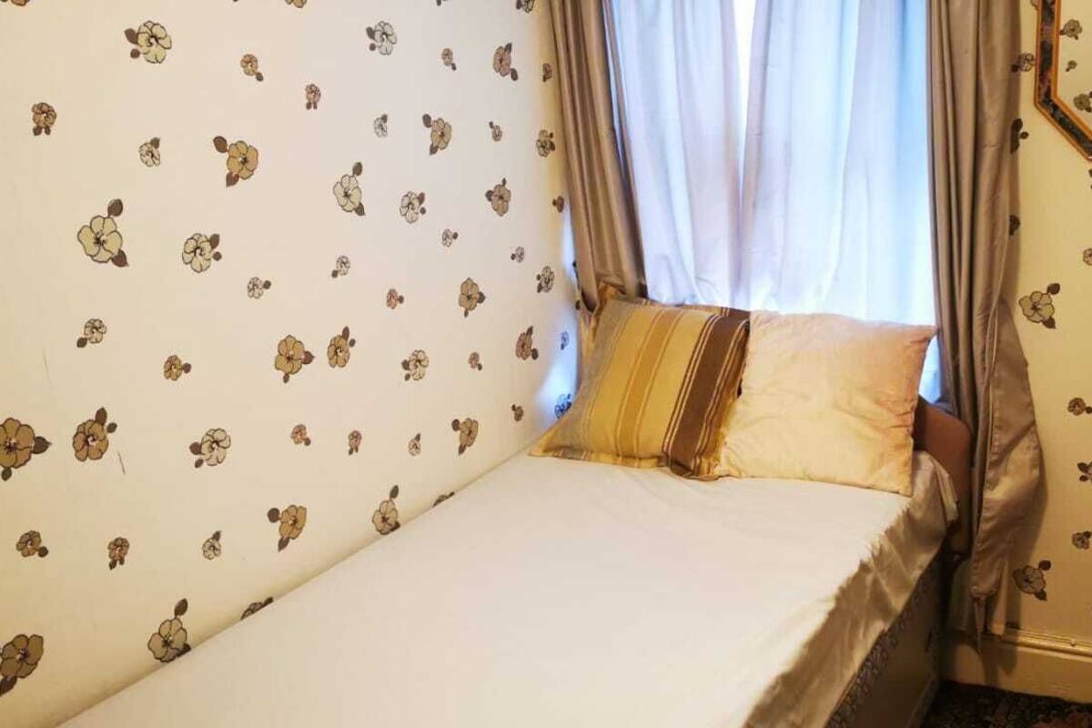 A small, single-bed room with floral wallpaper, a bedside table with a lamp, and a window with curtains.