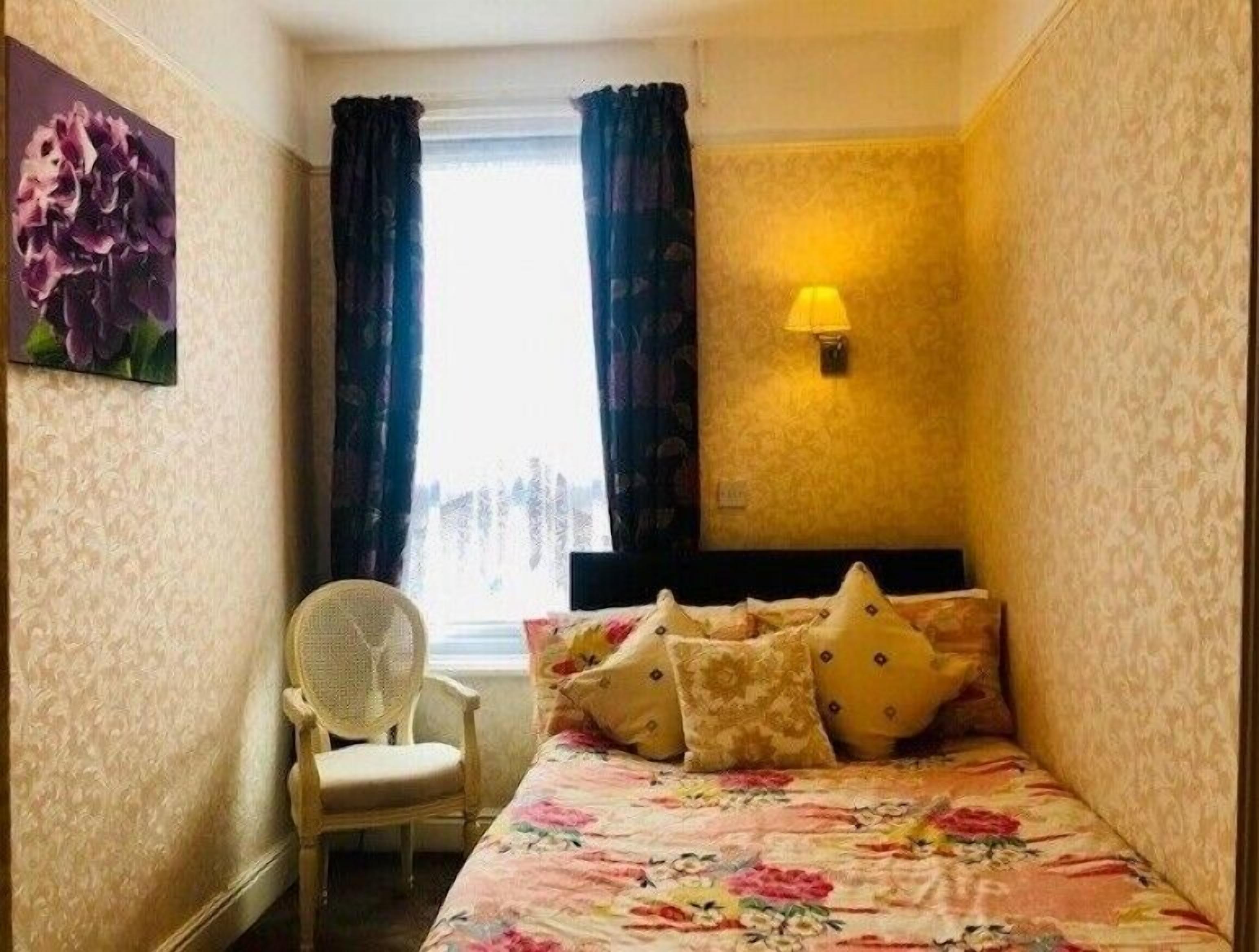 Una stanza piccola e accogliente con un letto, una sedia, una finestra con tende e una lampada a parete.