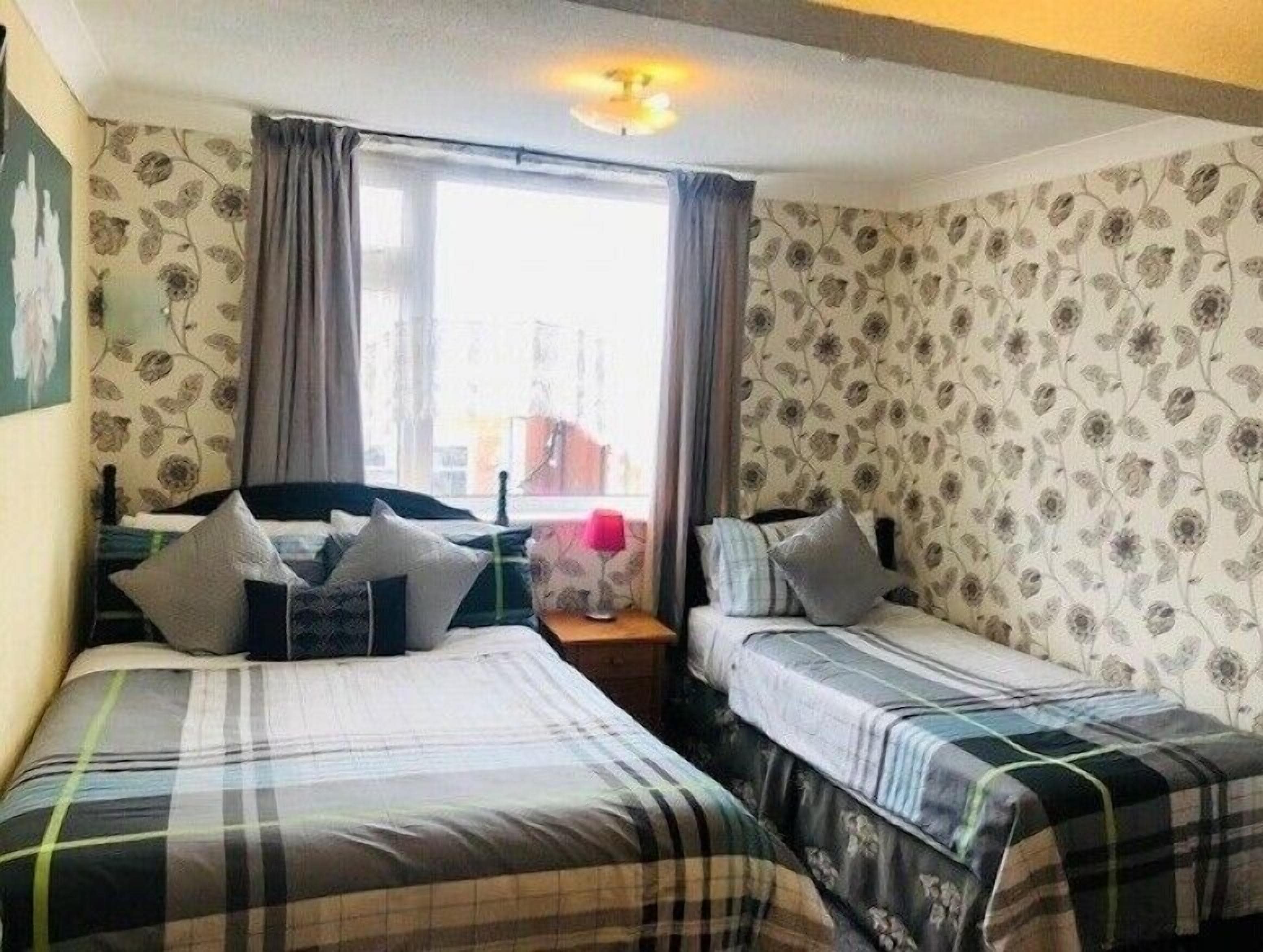 A room with two beds, a window with curtains, floral wallpaper, and a ceiling light.