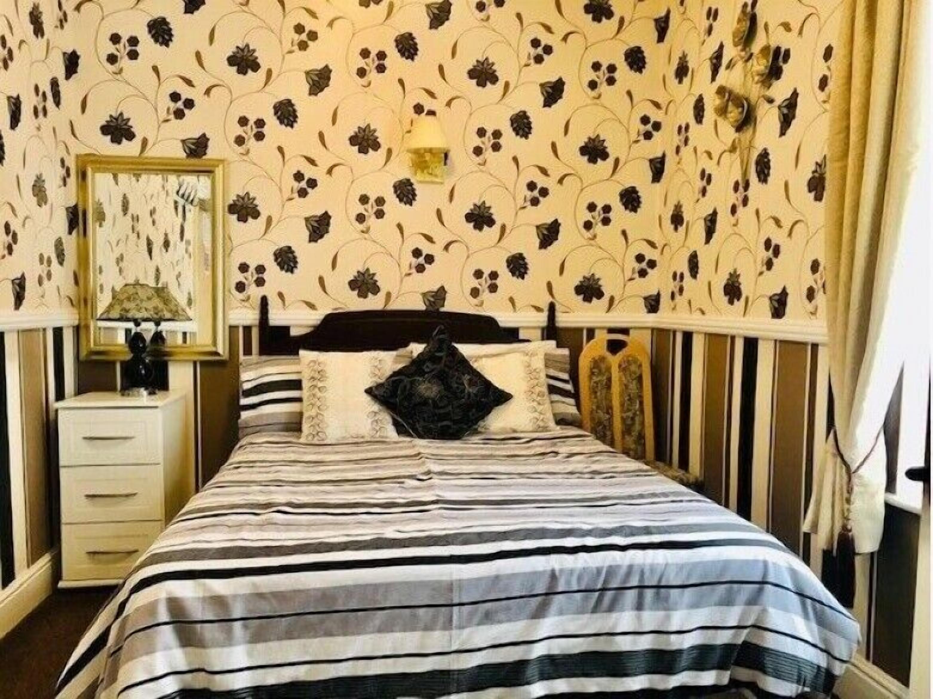 A bedroom with a striped bedspread, a floral wallpaper, a bedside table with a lamp, and a mirror.