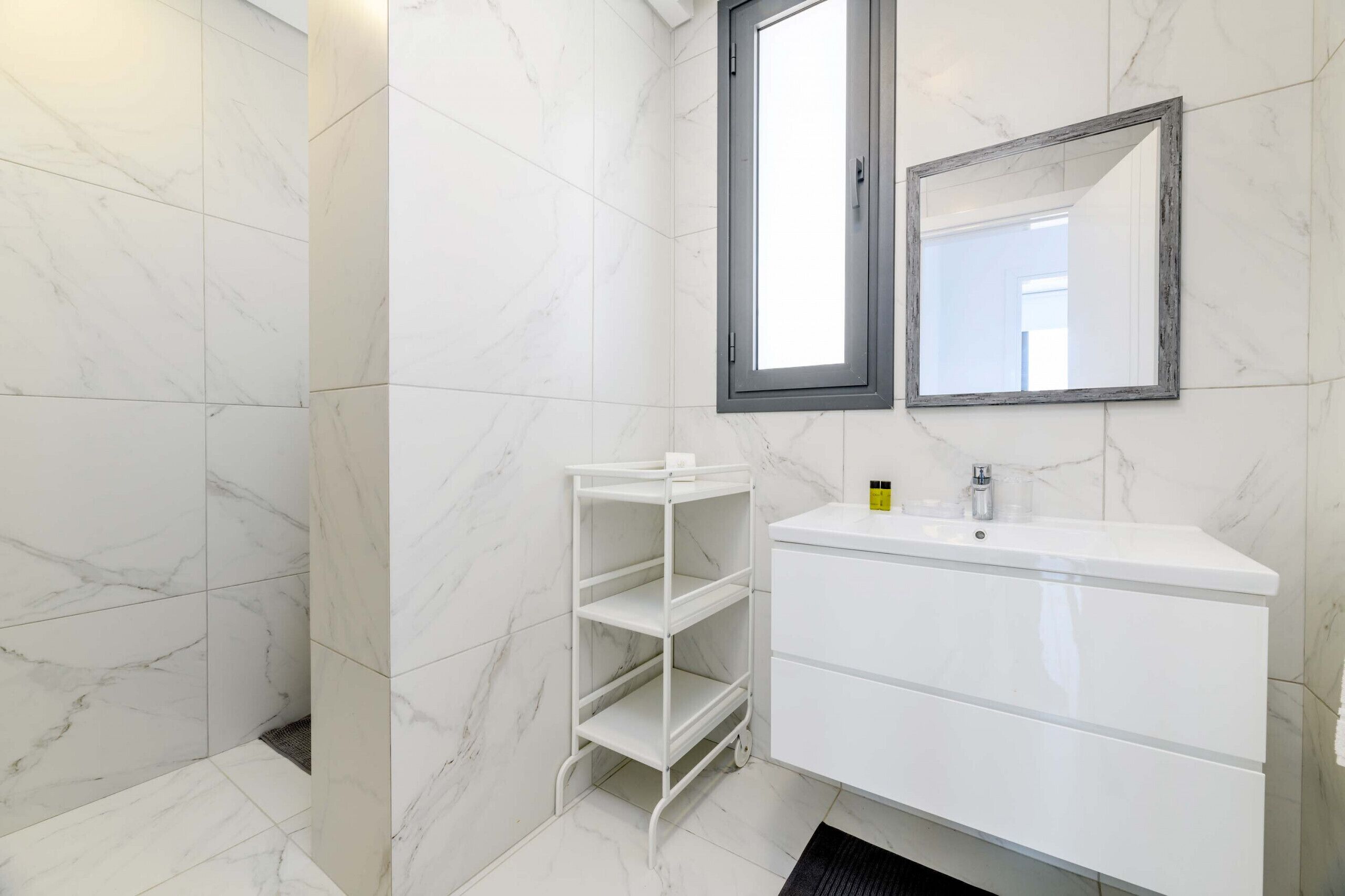 Une salle de bain avec des carreaux de marbre blanc, un meuble-lavabo blanc doté d’un miroir et une étagère métallique blanche.
