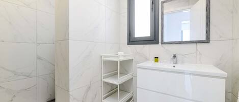 Une salle de bain avec des carreaux de marbre blanc, un meuble-lavabo blanc doté d’un miroir et une étagère métallique blanche.