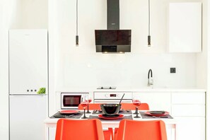 Eine moderne Küche mit weißen Schränken, einer schwarzen Dunstabzugshaube und einem Essbereich mit orangefarbenen Stühlen und einem weißen Tisch.