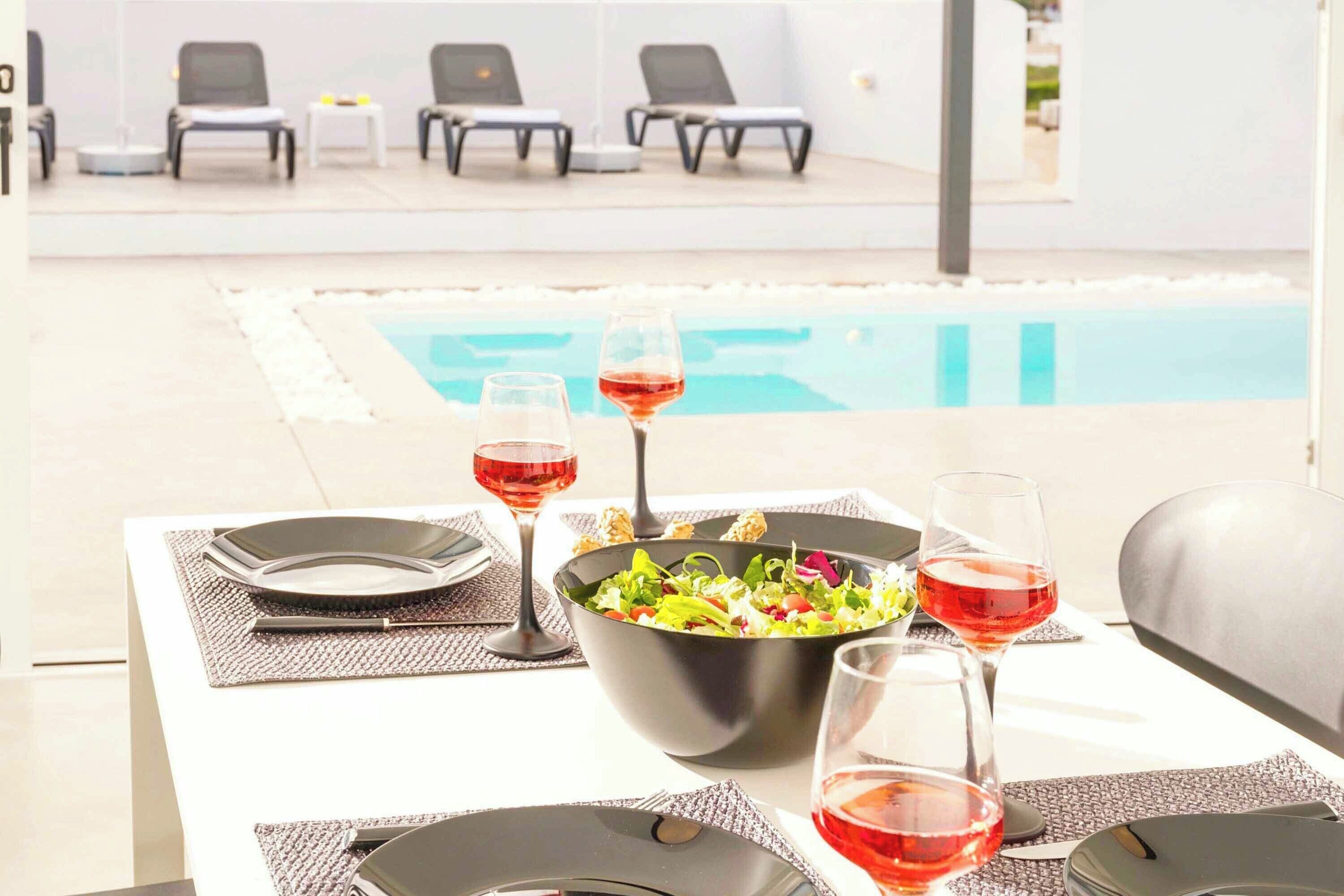 A dining table with a salad, wine glasses, and plates, overlooking a pool area with lounge chairs.
