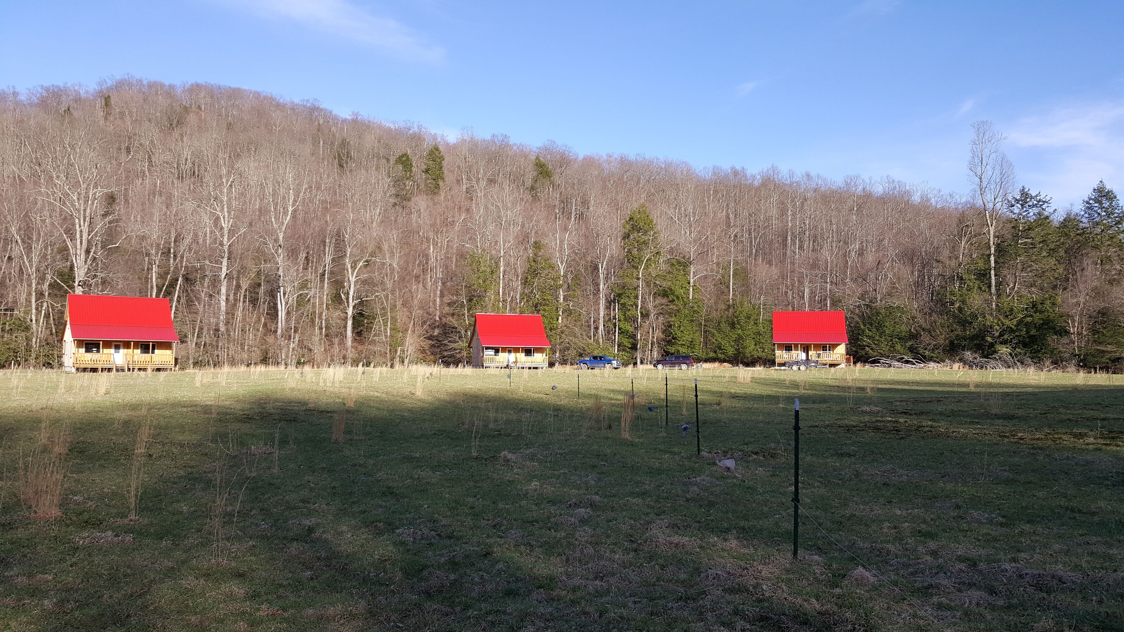 Deux cabanes aux toits rouges se dressent dans un champ verdoyant, entourées d’arbres et avec une colline en arrière-plan.