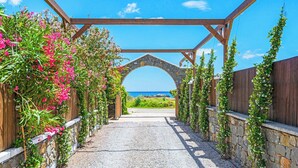 Ein Kiesweg führt zu einem steinernen Torbogen unter einer Holzpergola, gesäumt von blühenden Pflanzen und einem Holzzaun.