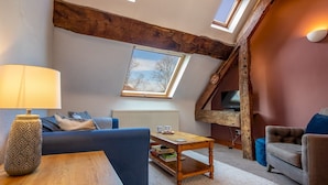 Een gezellige woonkamer met een hoge nok, zichtbare houten balken, een open haard en een televisie.