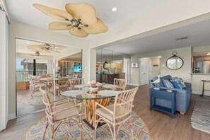 A spacious living area with a dining table, chairs, a blue sofa, and a ceiling fan. - Mariner Pointe 813: Top Floor Unit w/ Water Views! (Sanibel)
