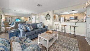 A modern living room with a grey sofa, a coffee table, and a kitchen area with a microwave and bar stools.