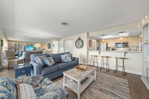 A modern living room with a grey sofa, a coffee table, and a kitchen area with a microwave and bar stools. - Mariner Pointe 813: Top Floor Unit w/ Water Views! (Sanibel)