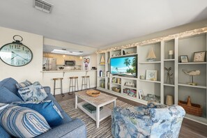 A living room with a sofa, coffee table, TV, and a kitchen in the background. - Mariner Pointe 813: Top Floor Unit w/ Water Views! (Sanibel)