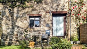 Una cabaña de piedra con una pequeña ventana, una puerta con cortina, un cartel que dice 'The Stables Cottage' y un jardín con plantas y una silla.