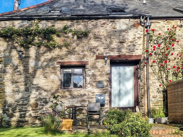 Ein Steinhäuschen mit kleinem Fenster, einer Tür mit Vorhang, einem Schild mit der Aufschrift 'The Stables Cottage' und einem Garten mit Pflanzen und einem Stuhl.