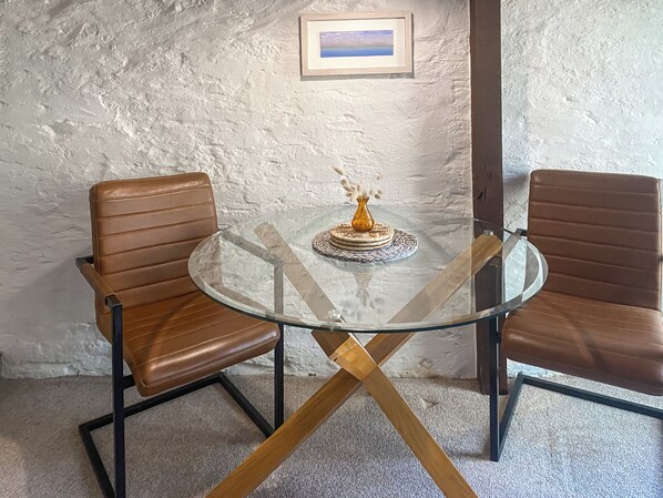 A room with a round glass table, two leather chairs, a framed picture on the wall, and a textured white wall.