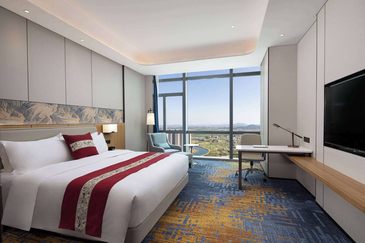 A hotel room with a large bed, a desk, a chair, a TV, and a view of the city.