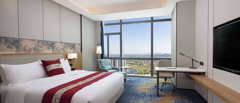 A hotel room with a large bed, a desk, a chair, a TV, and a view of the city.