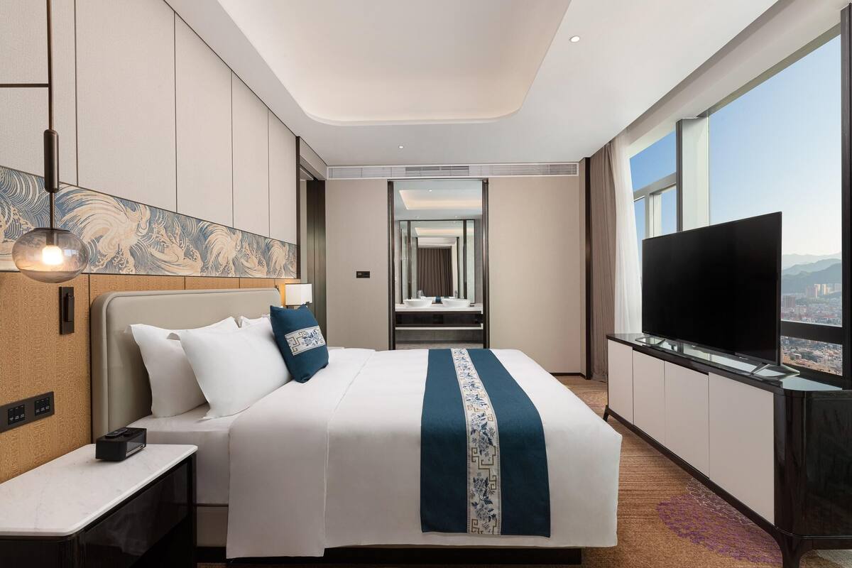 A hotel room with a large bed, a TV, a desk, and a view of the city.