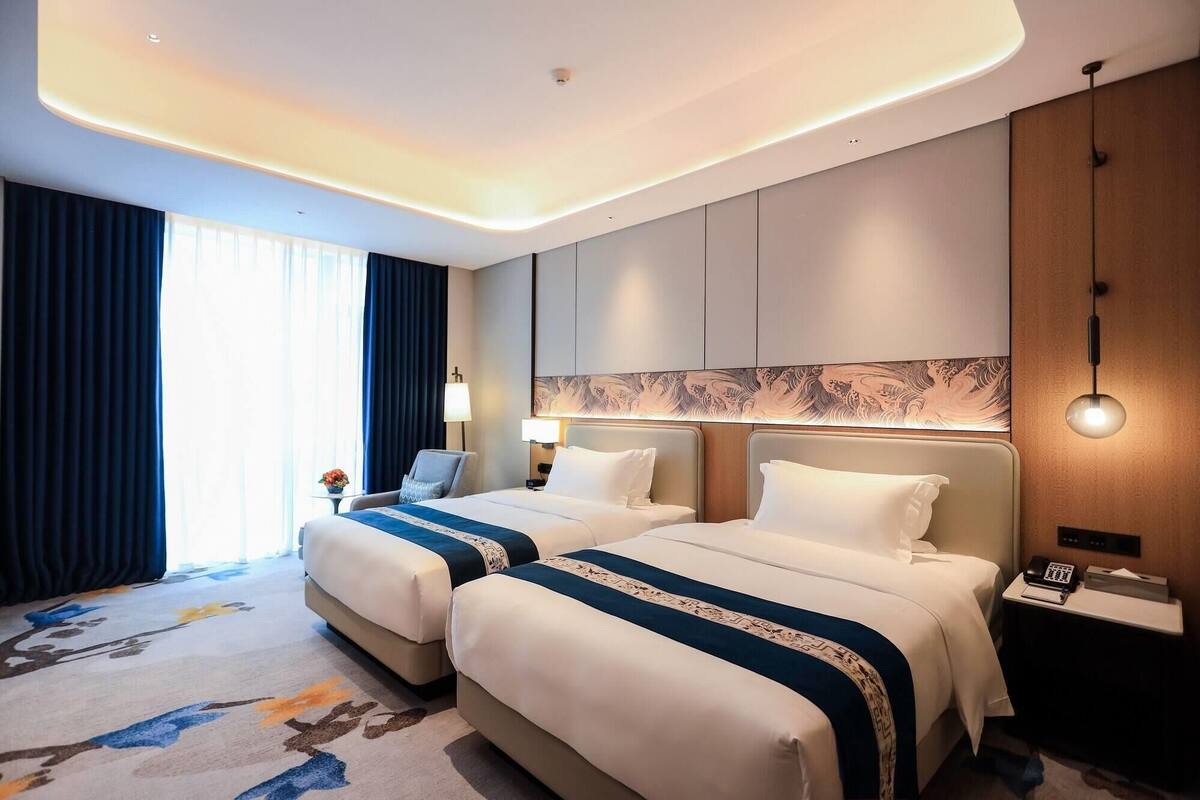 A hotel room with two beds, a desk, a lamp, and a view of the outdoors.