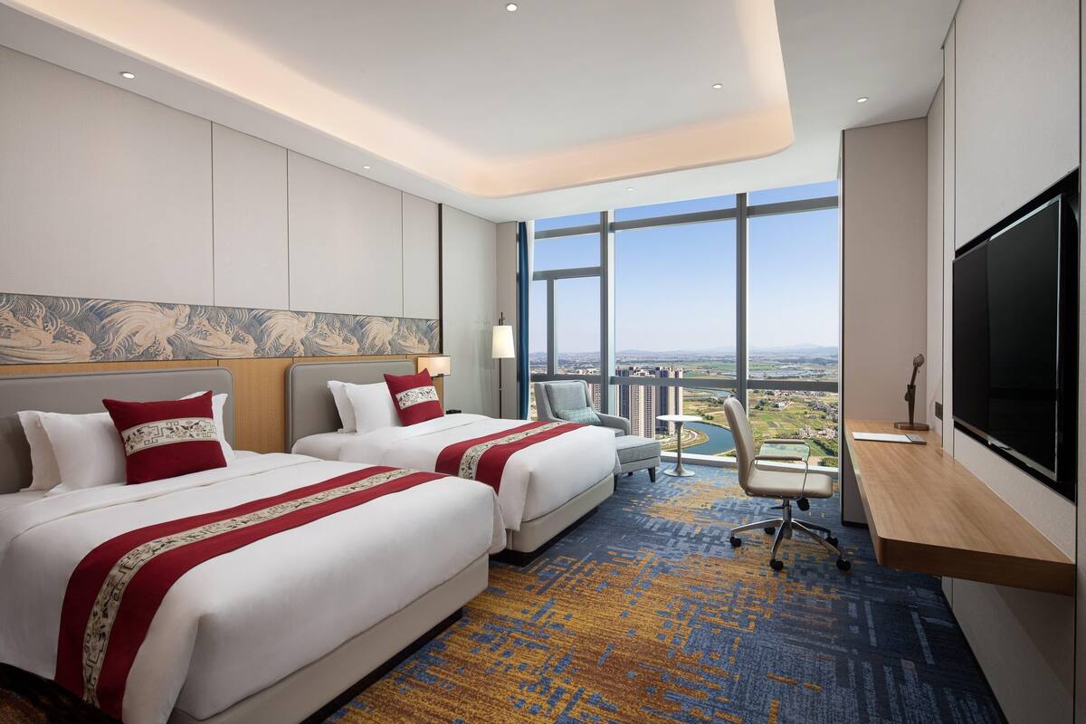 A hotel room with two beds, a desk, a chair, a TV, and a large window with a city view.