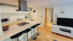 Une cuisine moderne avec un îlot central, des tabourets de bar, une cuisinière et un téléviseur posé sur un meuble blanc.