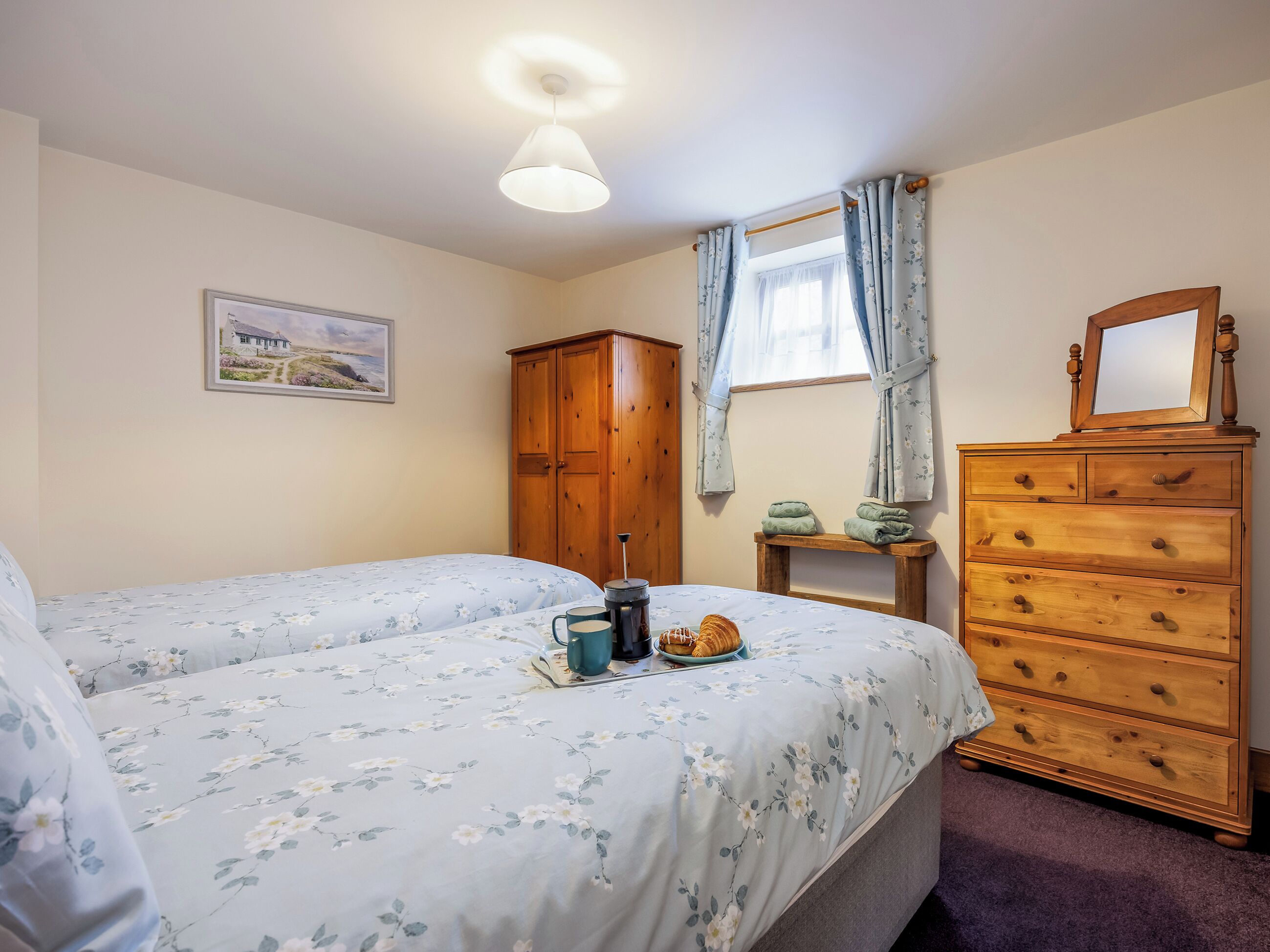 Une chambre avec deux lits, une armoire en bois, une petite table avec une bouilloire et des croissants, une fenêtre avec des rideaux et un tableau encadré au mur.