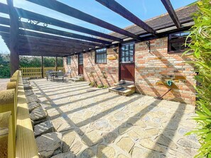 A covered patio area with stone flooring, wooden furniture, and a brick building with a wooden door.