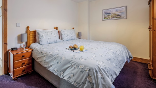 Quarto com cama, mesa de cabeceira de madeira e uma mesinha de apoio com abajur.