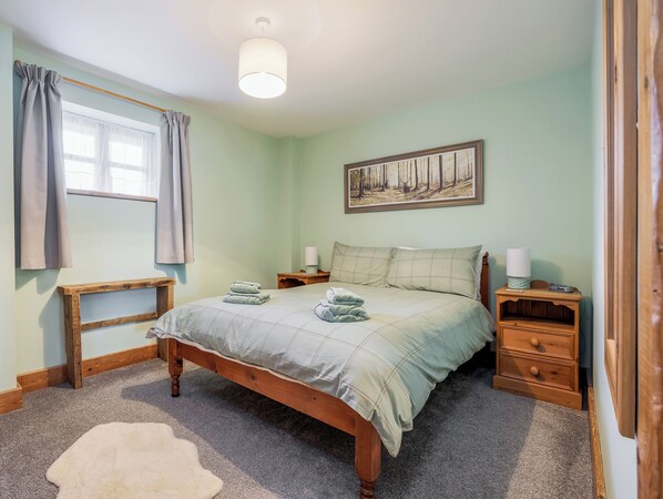 Ein Schlafzimmer mit einem hölzernen Bett, einem Nachttisch mit Lampe, einem Fenster mit Vorhängen und einem Bild an der Wand.