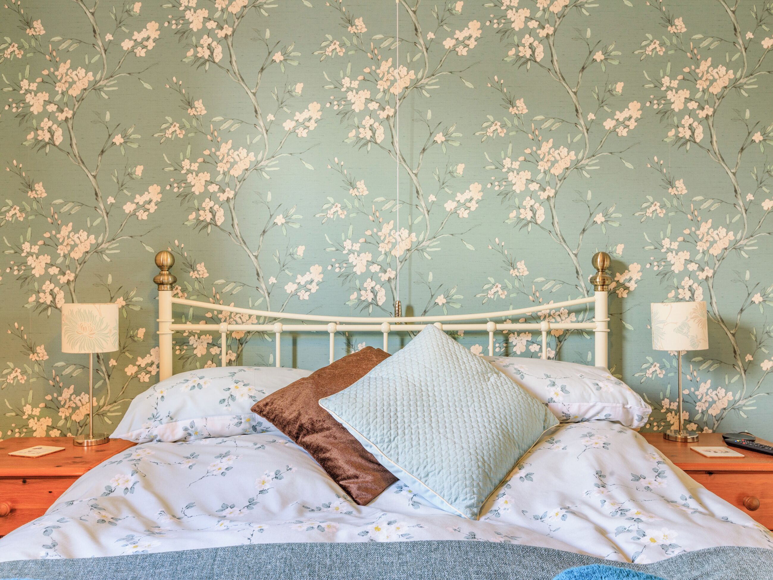 Un dormitorio con papel tapiz floral, un marco de cama blanco y dos lámparas de noche.