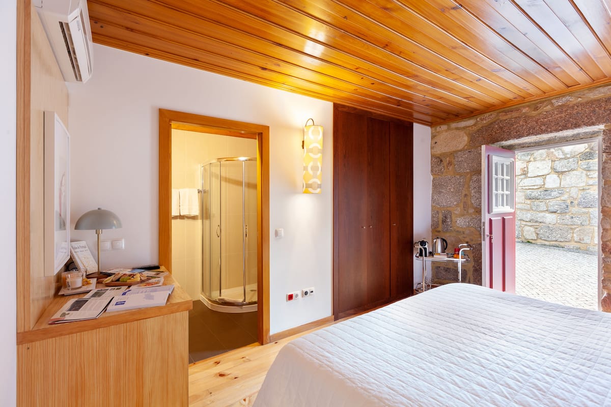 A hotel room with a wooden ceiling, a bed, a desk with a lamp, a shower, and a stone wall.