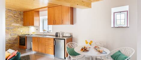 A compact kitchen with wooden cabinets, a built-in oven, and a sink. There is a dining area with white chairs and a table, and a small window with a view of greenery.