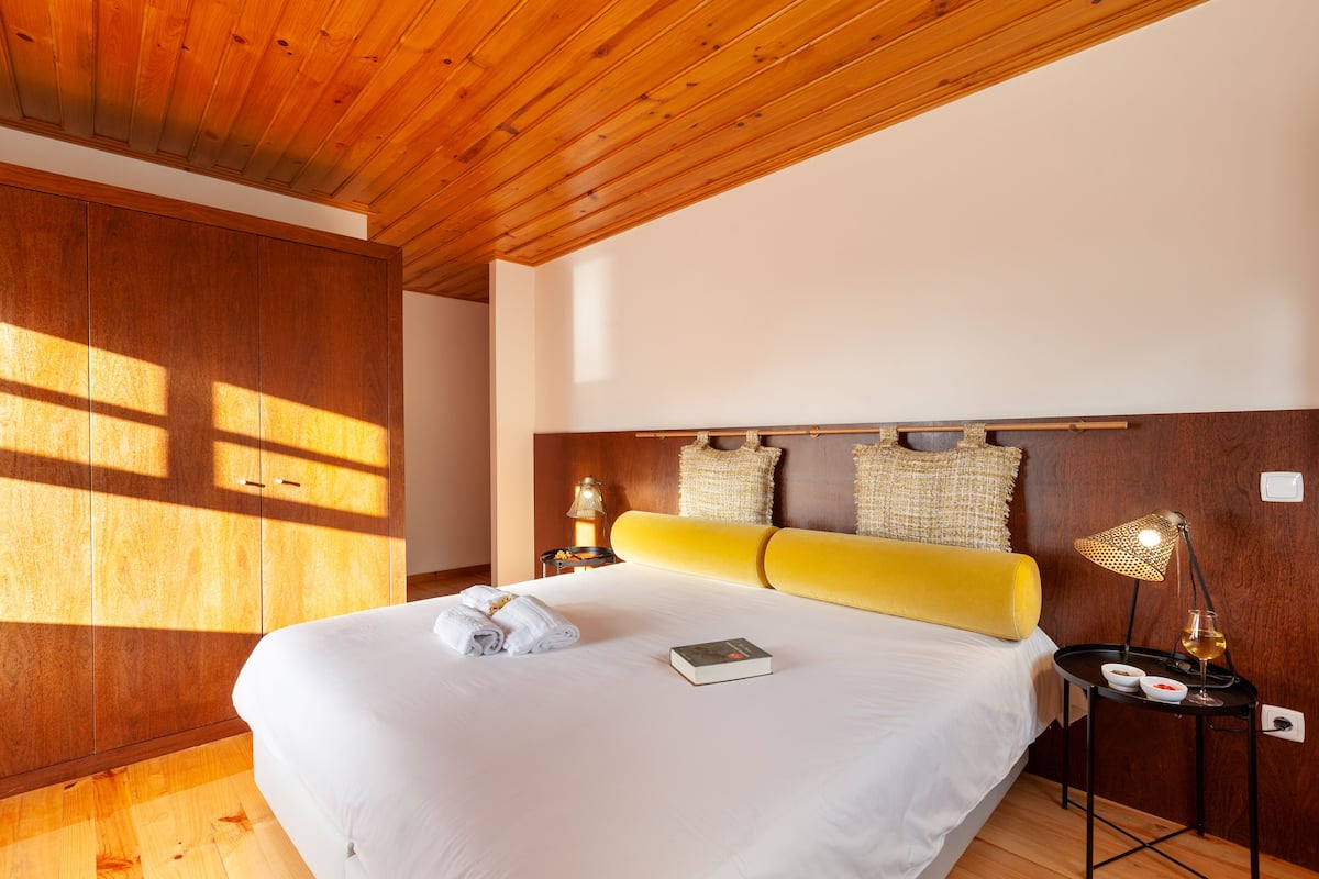 A hotel room with a wooden ceiling, a large bed with white bedding, a yellow headboard, and a bedside table with a lamp and a glass of wine.