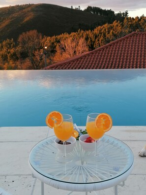 A glass table with two glasses of orange juice and a bowl of strawberries by a pool.