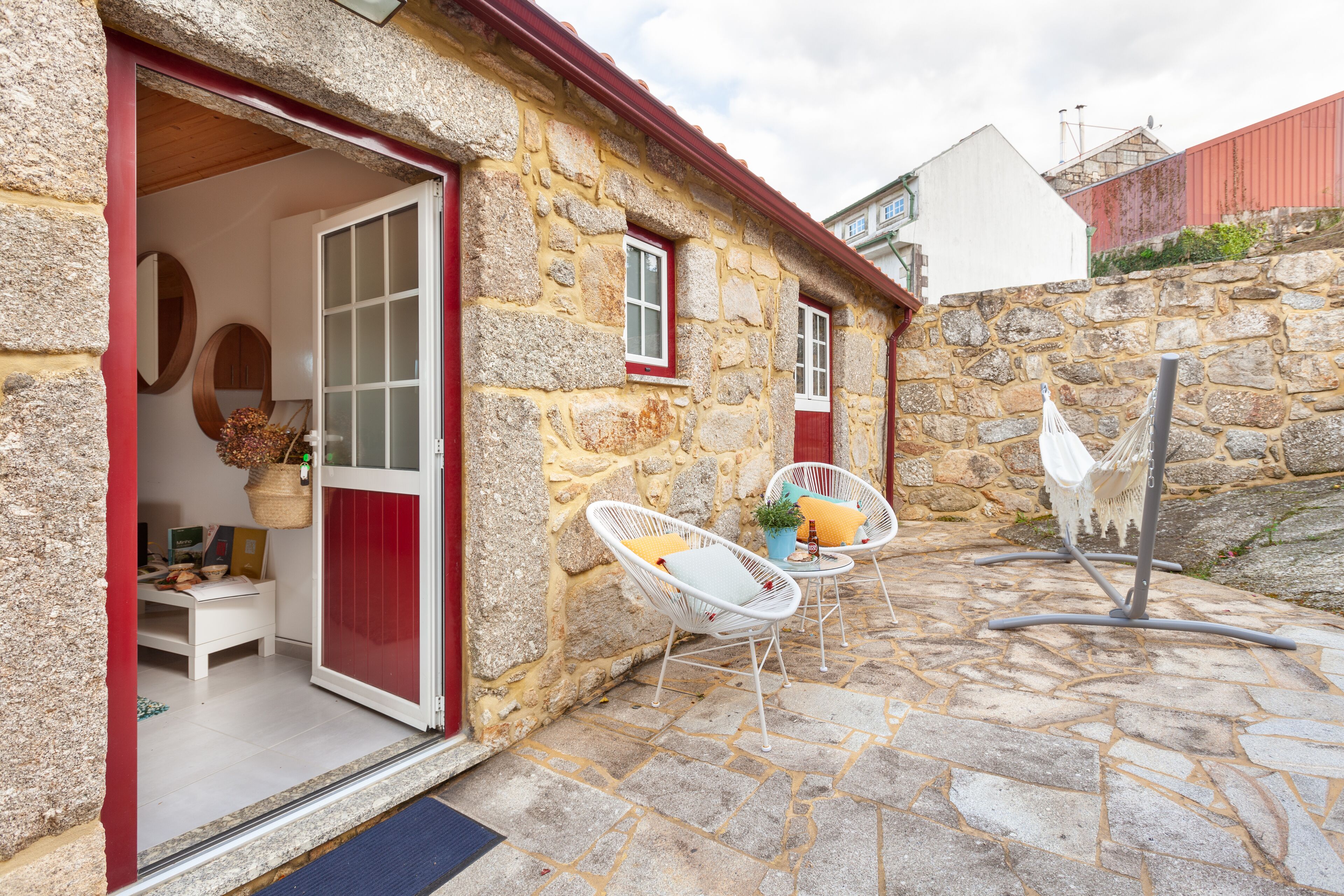 A stone building with a red door and white-framed windows. Inside, there is a white chair with a cushion and a small table with a plant.