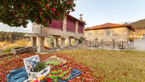 A stone building with a tiled roof, a patio with tables and chairs, and a picnic setup with a blanket, basket, and food. - CASA DO RANCHO (Amares)