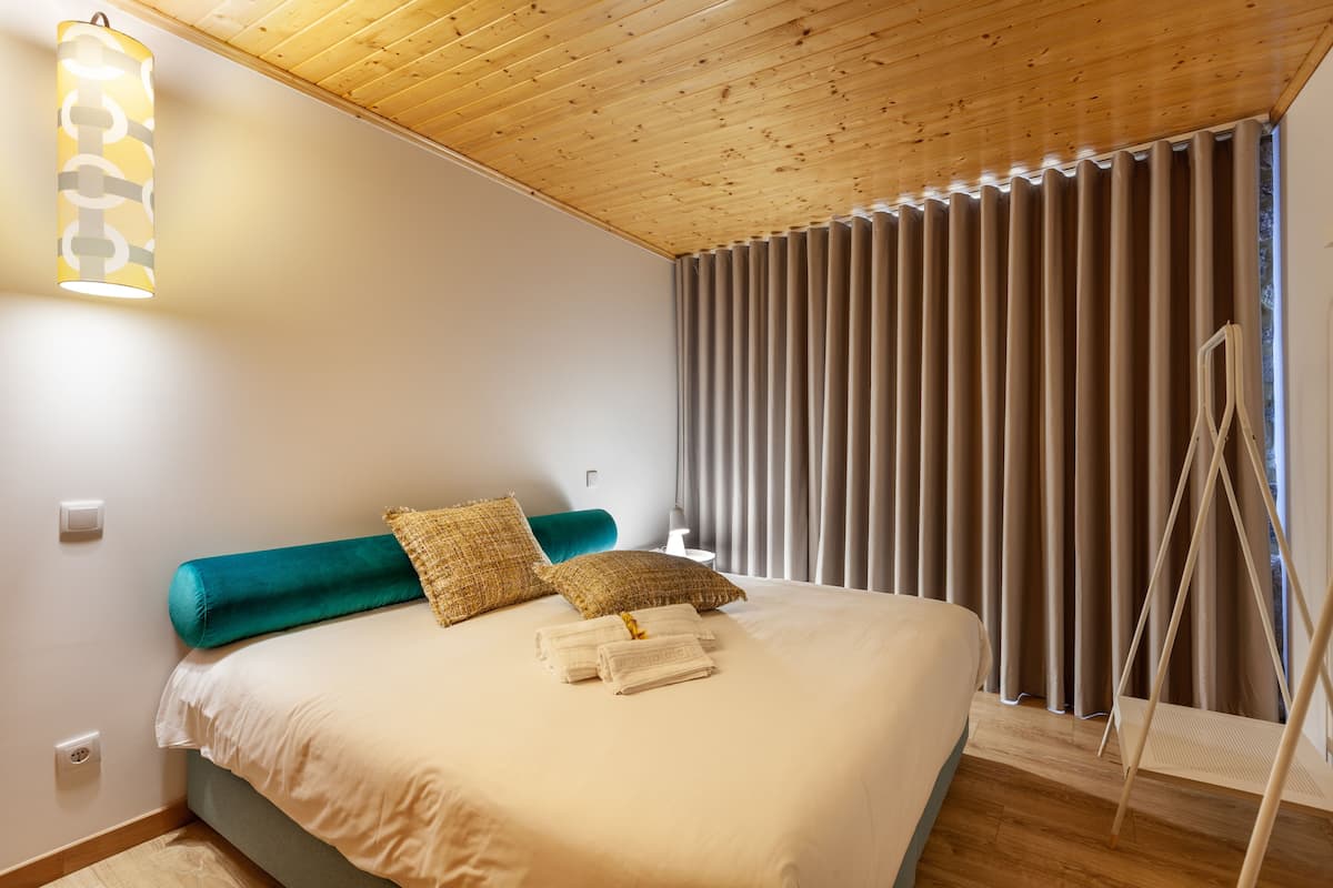 A neatly made bed with a teal headboard, two patterned pillows, and folded towels. A standing lamp is on the left, and a window with vertical blinds is on the right.