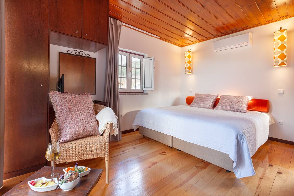 A bedroom with a bed, a wicker chair, a dining table with food, and a window.
